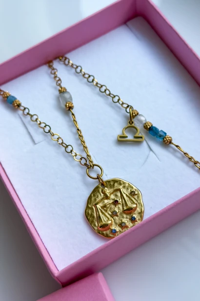 Naszyjnik dla zodiakalnej Wagi z kamieniem księżycowym i niebieskim akwamarynem. Złociste łańcuszki uzupełniają zawieszka ze znakiem Wagi oraz większy medalion z motywem wagi.