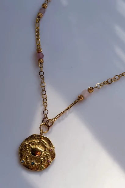 Detal naszyjnika Rak: medalion ze znakiem zodiaku, różowe kamienie i połączenie dwóch rodzajów złocistego łańcuszka.